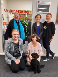 Five people and a dog pose outside an office window bearing the Volunteering Canterbury logo