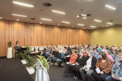 A large room full of seated people, looking towards the speaker, Glenda Martin, near the front stage