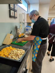 A man wearing a brightly coloured apron, cutting vegetables in a commercial kitchen. A tray of food on the bench in the foreground.