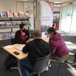 Three people sit around a table in a library. Shelves of magazines in the background. Volunteering Canterbury banner behind them