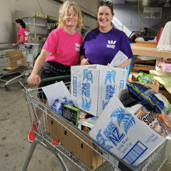 Two women from Westpac, standing in the 0800 Hungry warehouse with a trolley full of boxes and groceries