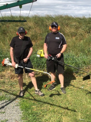 Two men standing in a grassy area, wearing ear muffs and holding weed whackers