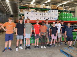 Students stand in a line in a food warehouse, in front of large shelves containing food and grocery items