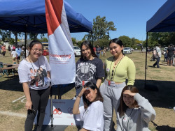 Three high school student volunteers stand near a Volunteering Canterbury flag in a park area. Two gazebos on either side of them