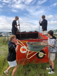 Four students at an orange rubbish skip. Two students stand in the skip holding spades, two students stand in front of the skip lifting a wheelbarrow