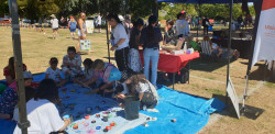 A group of adults and children sit on the ground painting small rocks. People stand at an information table behind them. Crowds of people milling in the background