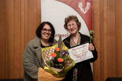 Tricia De Haan and a supporter, with her Award and a bunch of flowers, in front of the Volunteering Canterbury banner