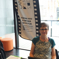 A women sitting at a table. The Christchurch Film Society banner in the background