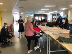 A woman stands in the centre of a large open school room. Students stand and sit around looking towards her. A table is strewn with pieces of fabric