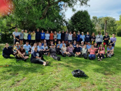 A very large group of people stand and sit in the shade. A variety of trees in the background