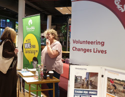 A woman stands behind a stall, talking to a student about volunteering. Volunteering Canterbury banner in the foreground. Tourettes Association of NZ banner in the background