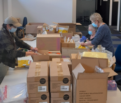 A large table covered with boxes. Three women stand and sit around the table, wearing masks and packing contents into boxes