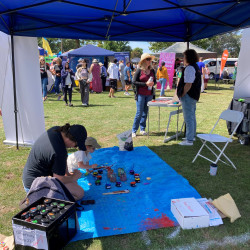 The Volunteering Canterbury stand at Culture Galore. Children sitting on a blue mat painting rocks in the foreground. Two women speaking about volunteering in the background