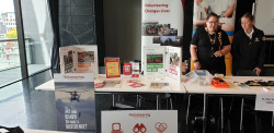 A table displaying Volunteering Canterbury information pamphlets, posters and banner. To the right, two people stand behind a table with a sign saying "St John Youth Cadets"