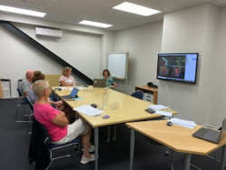 People sitting around a table in a meeting room. A TV on the wall displays participants on a Zoom meeting