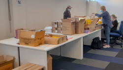 A large table covered with boxes. Three women stand and sit around the table, wearing masks and packing contents into boxes