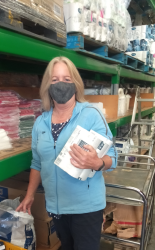 A woman wearing a face mask stands in front of large shelves, holding items to be packed into a food box