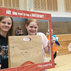 Two young girls looking through the red Volunteering Canterbury frame, holding a decorated brown paper bag