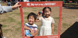 Two children hold up their painted rocks for the camera, as they look through the red cardboard sign that reads "Are you ready to make a difference?