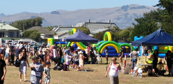 A large crowd of people in a park. An inflatable obstacle course in the background and the Christchurch Porthills behind them