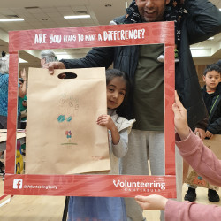A father with his two young children, looking through the red Volunteering Canterbury frame, holding a decorated brown paper bag