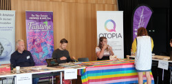 People sitting at display tables. Banners for Christchurch Amateur Radio Club, The NZ Comics and Art Zine, and Qtopia in the background