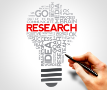 A collection of printed grey words in the shape of a lightbulb. The central word in red reads "RESEARCH". A hand holds a pen ready to write.