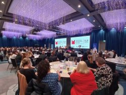 A wide-angle view of the conference room, showing attendees sitting at round tables, with the stage and screen in the background, and purple decorative lights hanging from the ceiling