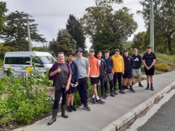 A woman and nine Christs' College students stand in a line in front of a garden bed of sunflowers. A school van is parked behind them. Trees and bushes in the background