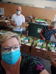 In the foreground, a woman with a face mask takes a selfie. Behind her is a table of boxes and two people packing contents into the boxes