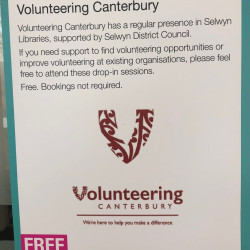 A sign in the library that reads "Volunteering Canterbury has a regular presence in Selwyn Libraries, supported by Selwyn District Council. If you need support to find volunteering opportunities or improve volunteering at existing organisations, please feel free to attend these drop-in sessions. Free. Bookings not required."