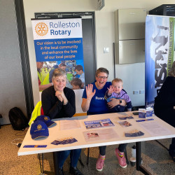 Two adults and a baby sitting behind a table, with the Rolleston Rotary flag
