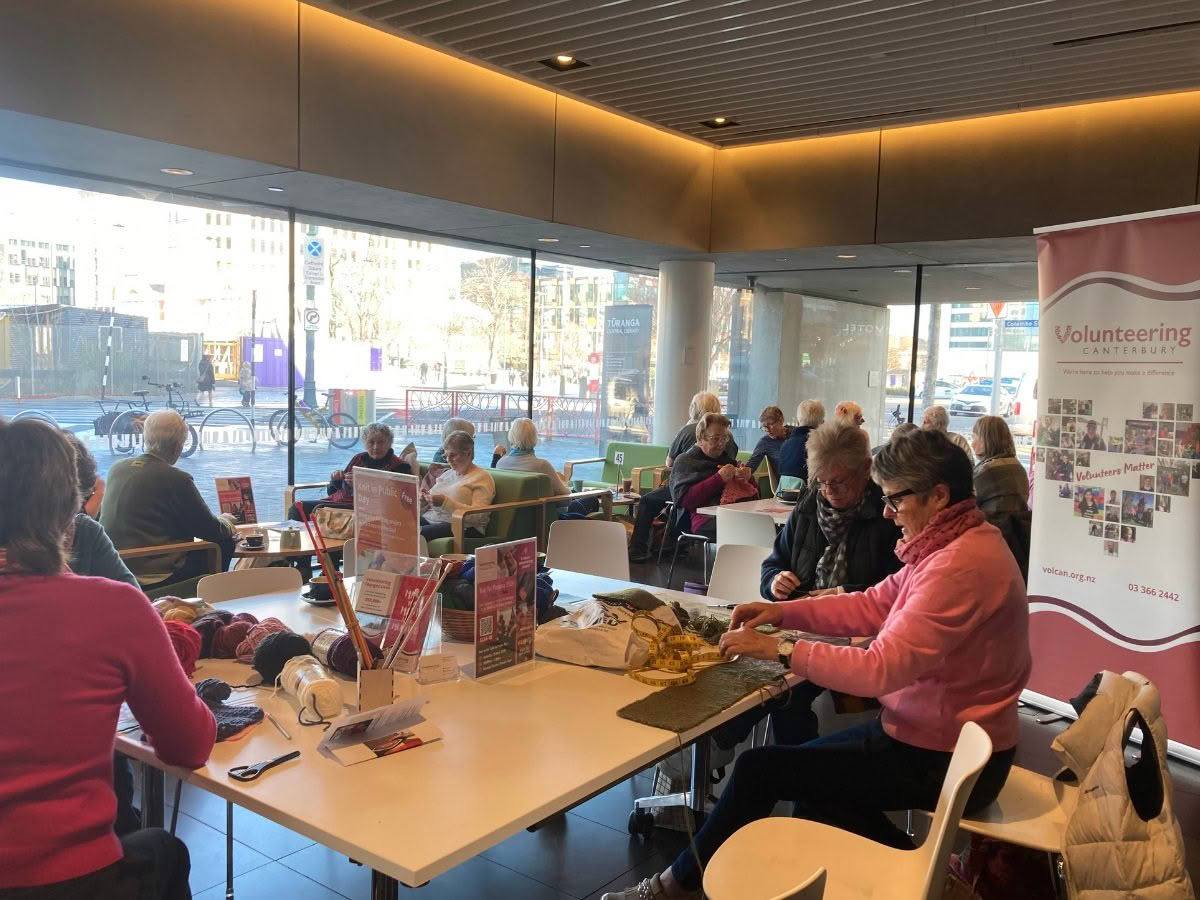 A large room full of people seated at tables, knitting in public together.