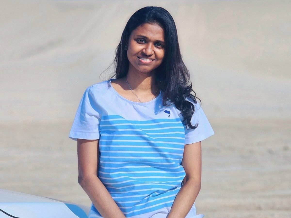 An image of a smiling young woman, perched on the front of a car bonnet, in front of a sandy-coloured background.