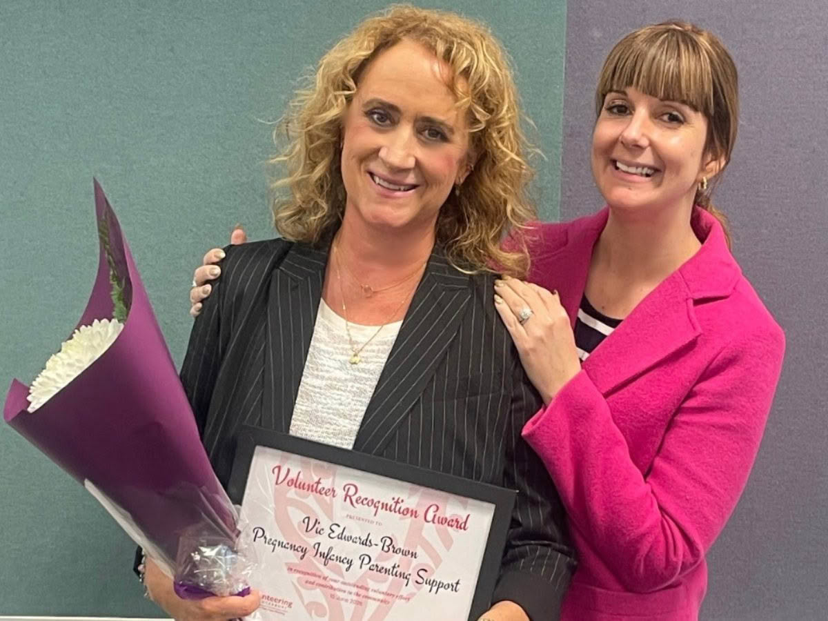PIPS Volunteer, Vic, with a friend, smiling at the camera and holding her Volunteer Recognition Award and a bunch of flowers.